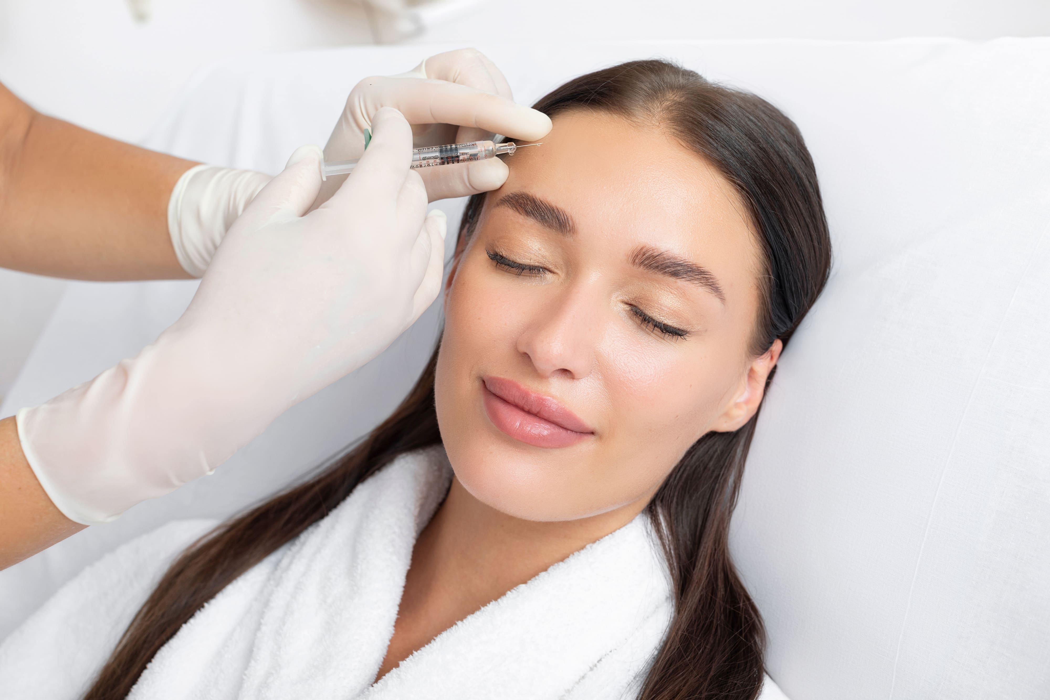 Gloved hands injecting a neuromodulator above the brow of a smiling patient lying on a treatment couch